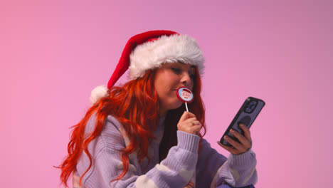 Studio-Shot-Of-Young-Gen-Z-Woman-Wearing-Christmas-Santa-Hat-Eating-Candy-Lollipop-Looking-At-Mobile-Phone